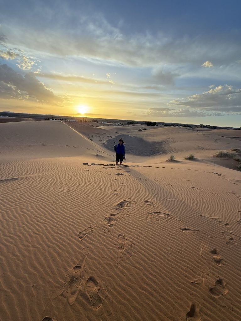 Randonneur marchant dans le désert de M’Hamid au coucher de soleil avec dunes dorées en arrière-plan.